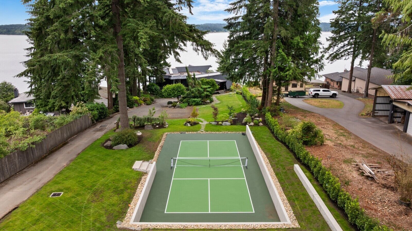 A tennis court surrounded by a fenced yard with a view of a lake and residential buildings.