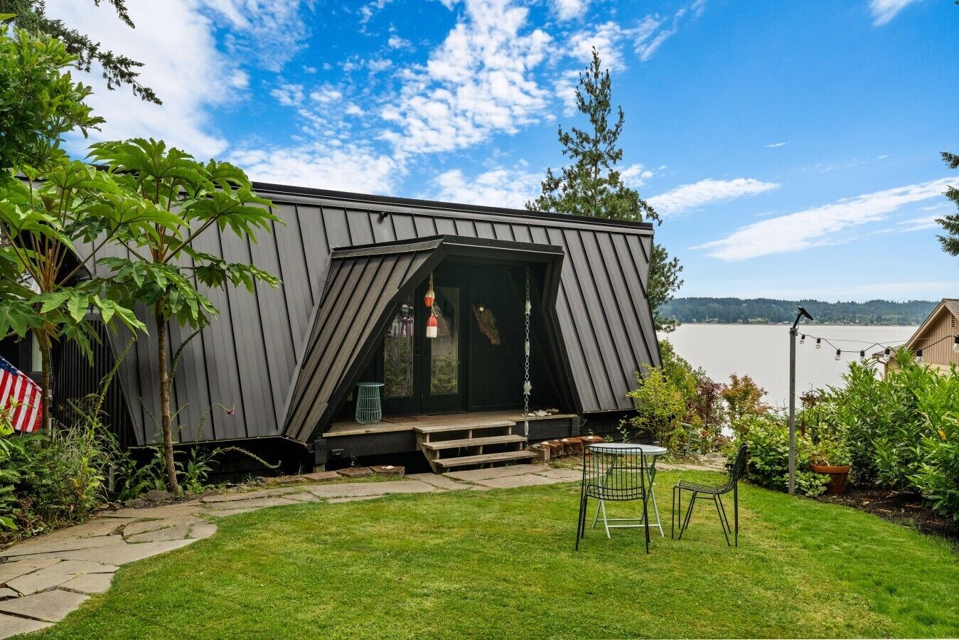 A modern houseboat with a deck, a small table, and chairs, surrounded by greenery and a body of water in the background.