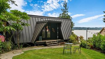 A modern houseboat with a deck, a small table, and chairs, surrounded by greenery and a body of water in the background.
