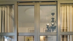 Een kamer met een groot raam, verdeeld in vier delen, elk met een metalen lijst. Het raam biedt uitzicht op het interieur. Er staat een bed met een metalen frame en hoofdbord, en een klein tafeltje met een stoel.