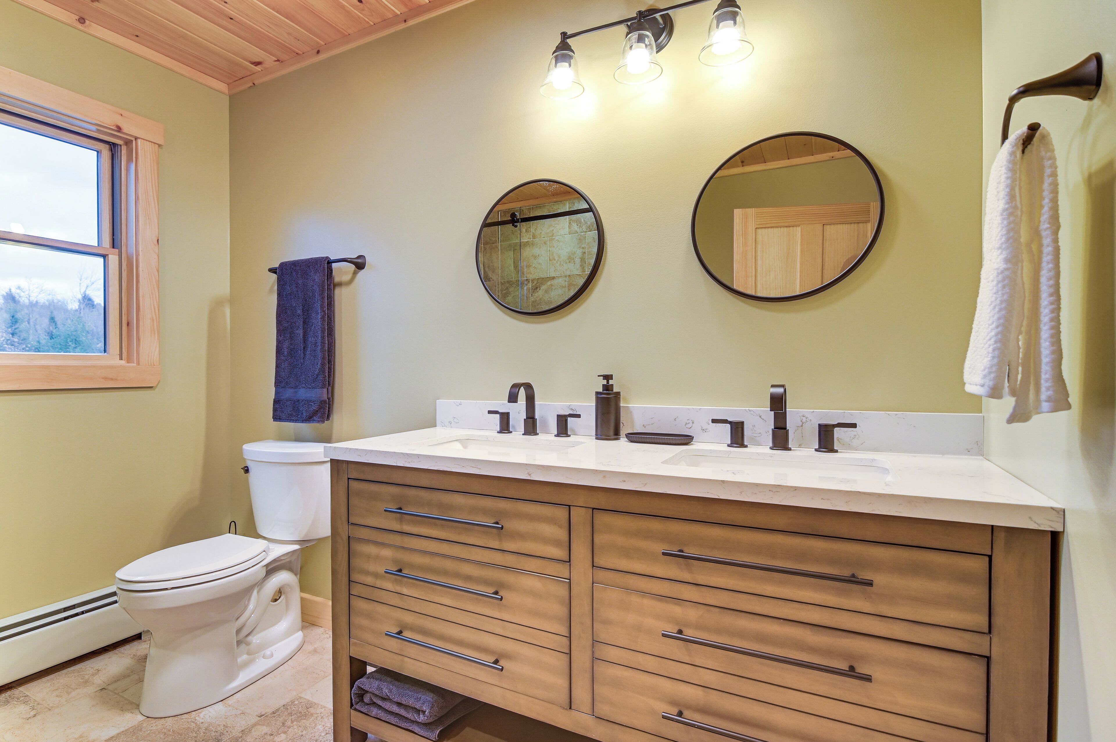 A bathroom with a white toilet, a double sink vanity with wooden cabinets, and two round mirrors.
