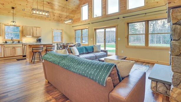 A spacious living area with a large brown sofa, a stone fireplace, and a view of the outdoors through large windows. - Hot Tub | Fire Pit | Mtn Escape Near Sunday River (Bethel)