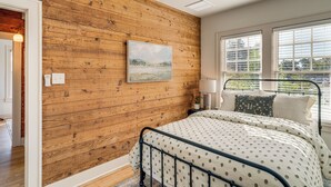 Une chambre avec un mur en bois, un lit recouvert d’une couvre-lit à motifs, une fenêtre avec des stores et un tableau accroché au mur.