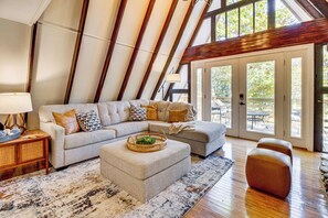 Um loft espaçoso com teto alto, vigas de madeira aparente, um amplo sofá de canto, um pequeno banquinho e uma área de refeições visível através de portas de vidro.