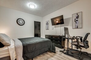 A bedroom with a bed, a desk with a computer, a chair, a TV, and artwork on the walls. - Easy Hwy Access to ATL: Work-Ready Riverdale Home (Riverdale)