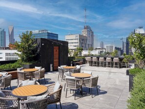 Un'area salotto all'aperto con tavoli rotondi e sedie in vimini, circondata da vegetazione e con vista su un paesaggio urbano caratterizzato da edifici moderni.