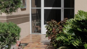 Une entrée fermée par un portail, agrémentée d’un panneau indiquant « RING », de plantes en pot et d’une terrasse carrelée.