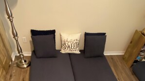 Une chambre avec un lit doté de draps sombres, d’une lampe et d’un coussin orné de la mention « chez soi, quel bonheur ».
