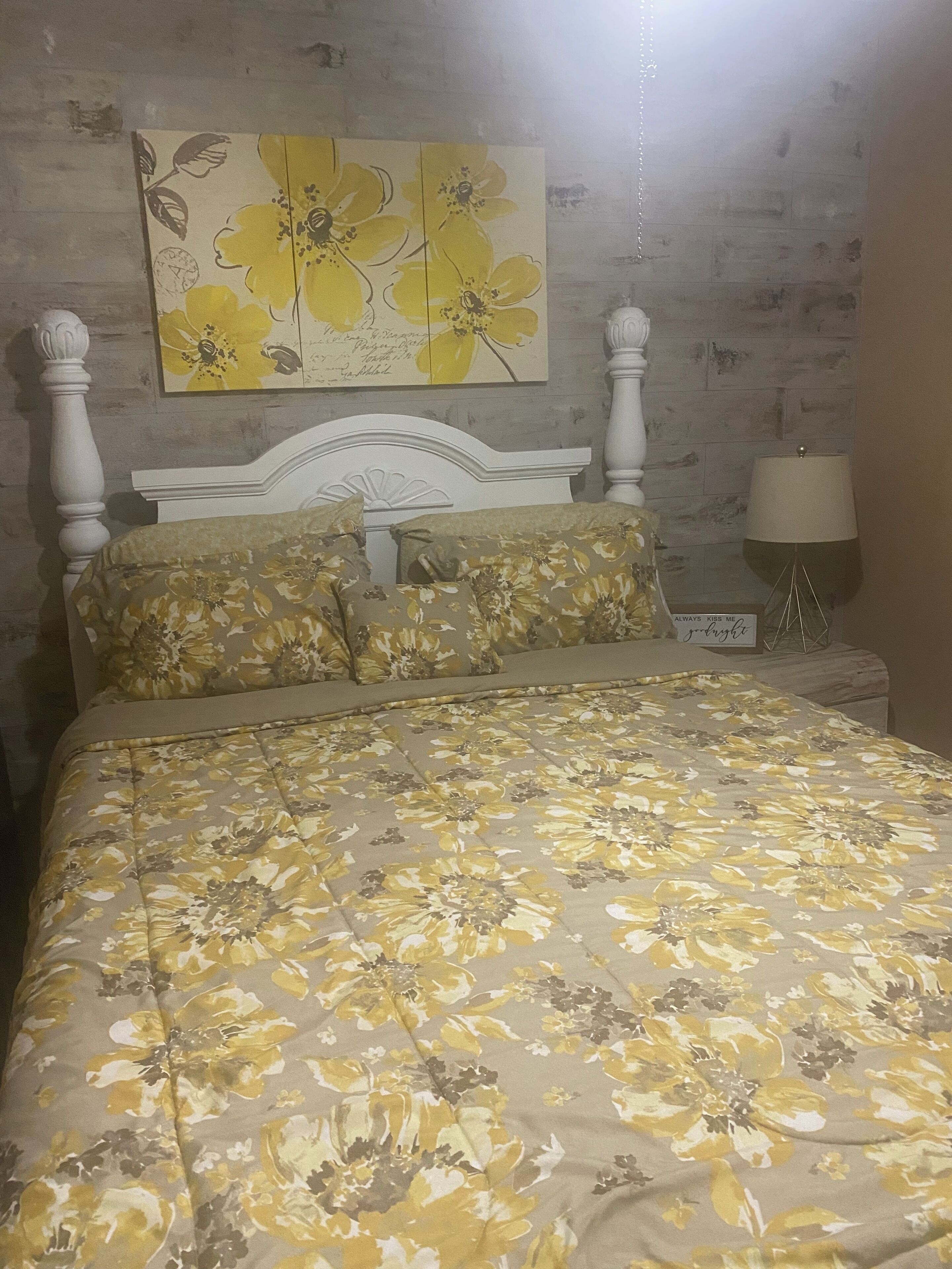 Un llit amb llençols florals, un panell de capçal blanc i un quadre de flors penjat a la paret.