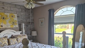 Une chambre à coucher comprenant un lit, un ventilateur de plafond, une fenêtre avec des rideaux, un tableau accroché au mur et une table de chevet.