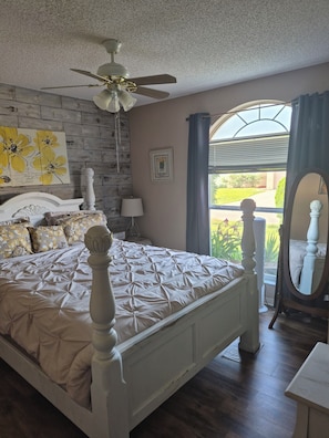 Une chambre à coucher comprenant un lit, un ventilateur de plafond, une fenêtre avec des rideaux, un tableau accroché au mur et une table de chevet.