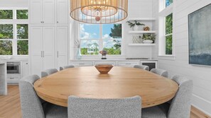 A spacious dining area with a large wooden table surrounded by grey upholstered chairs, white cabinetry, and a large window offering a view of greenery and a house. - Spacious Coastal Retreat Near Inlet Beach With Bikes, Balconies & Modern Design Bluewater View by Avantstay (Panama City Beach)