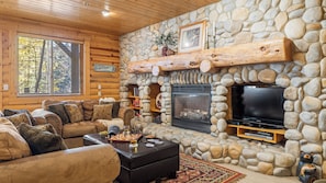 A log cabin interior with a stone fireplace, a television, a leather sofa, and a chessboard.
