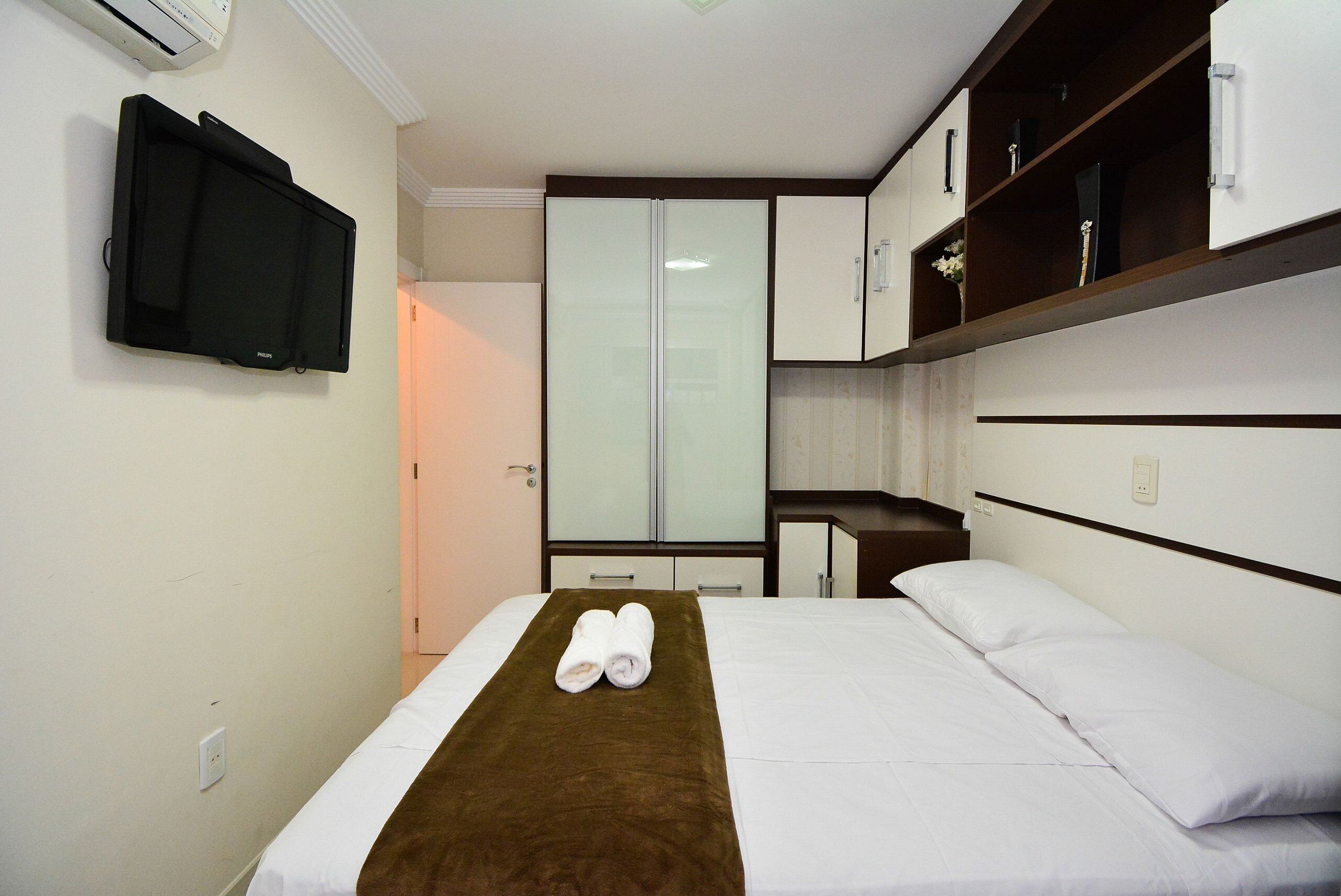 Habitación de hotel con cama, una manta marrón, televisor de pantalla plana montado en la pared, armario y un escritorio pequeño con una silla.