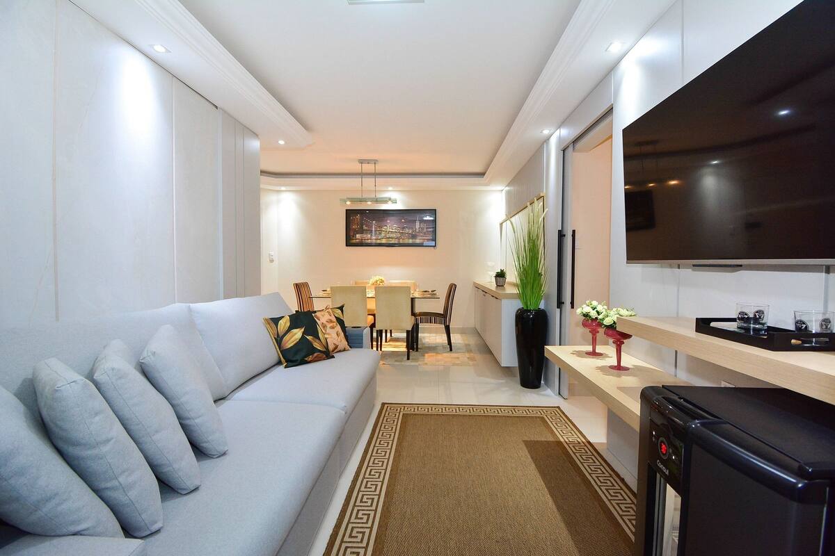 Una sala moderna con sofá, mesa de centro y televisión. Se observa un comedor con sillas y mesa al fondo.