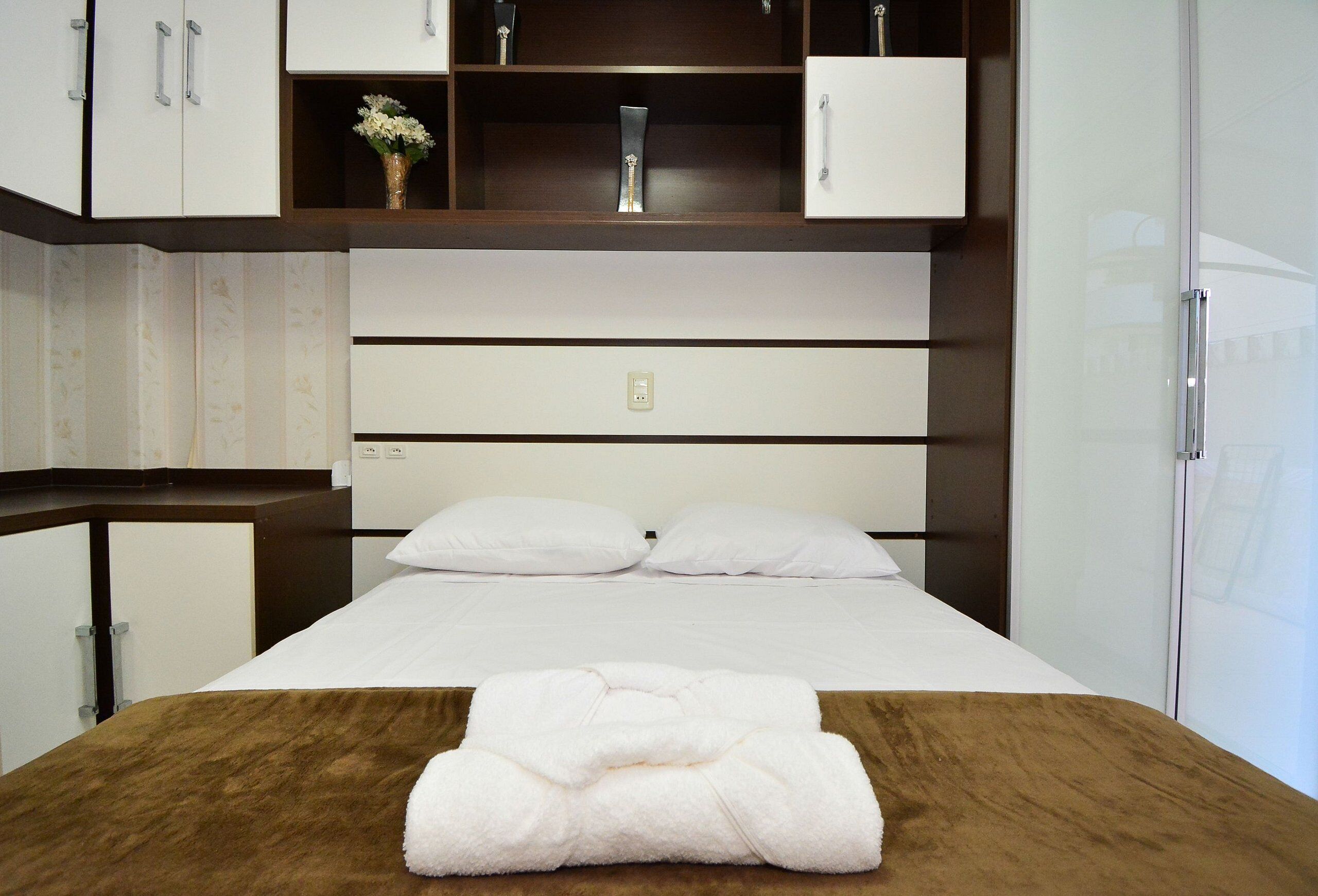 A hotel room with a bed, a wooden headboard, a white bedside table, a white towel, and a wardrobe with a mirror.