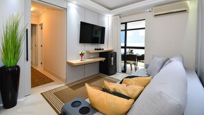 En moderne stue med en grå sofa, et fladskærms-tv, et køkkenområde med et køleskab, og et spiseområde kan ses igennem en glasdør.