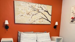 Un lit bien fait, avec une literie à rayures, deux tables de chevet avec des lampes, un tableau représentant un arbre accroché au mur, et une petite étagère avec des livres.