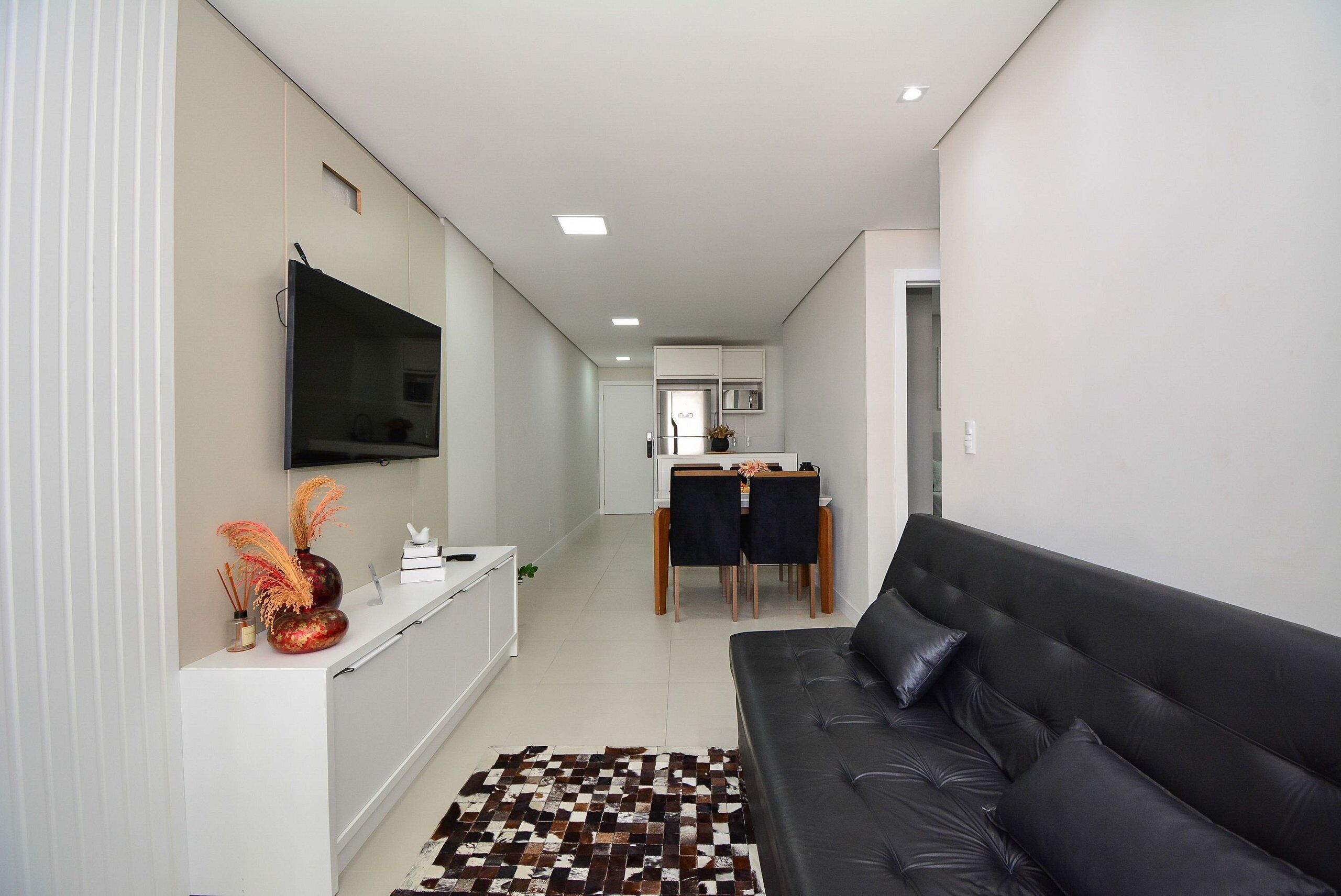 Un salon moderne avec un canapé en cuir noir, un téléviseur à écran plat fixé au mur et un coin repas comprenant une table et des chaises.