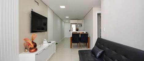 Un salon moderne avec un canapé en cuir noir, un téléviseur à écran plat fixé au mur et un coin repas comprenant une table et des chaises.