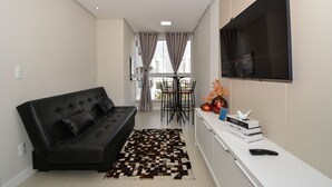 A modern living room with a black sofa, a patterned rug, a TV, and a small table with books and decorative items. - Apartamento Aconchegante em Itajaí (Itajaí)