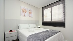 A bedroom with a bed, a nightstand, a lamp, and a window with blinds. - Apartamento Aconchegante em Itajaí (Itajaí)