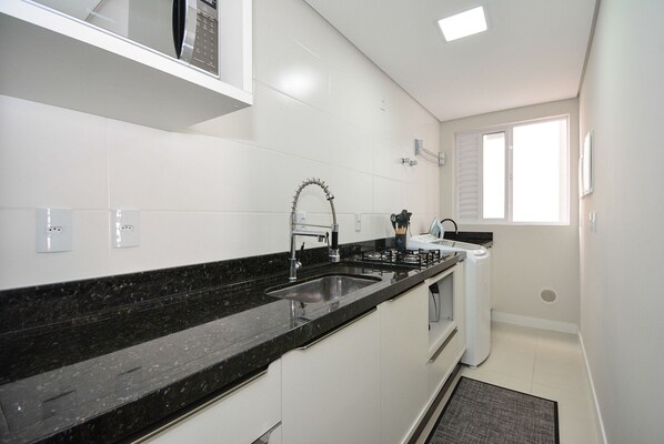 A kitchen with white cabinets, a black countertop, a stainless steel sink, and a window. - Apartamento Aconchegante em Itajaí (Itajaí)