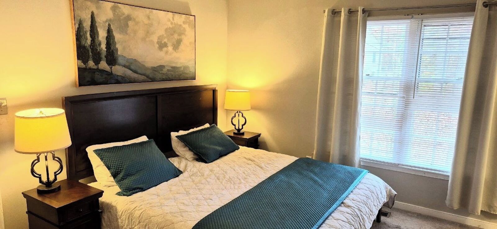 Una cama bien hecha con colcha, dos almohadas y cabecero. Dos lámparas de noche sobre mesitas de madera. Un cuadro cuelga sobre la cama, y se ve una ventana con cortinas.