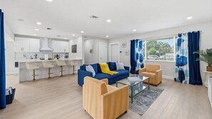 Ein modernes Wohnzimmer mit einem blauen Sofa, Holzsesseln, einem Glastisch und einer Küchenzeile mit weißen Schränken und Barhocker.