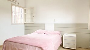 Een slaapkamer met een bed, nachtkastje, dressoir, een raam met tralies en een airconditioner.