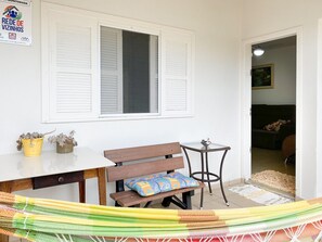 Una habitación con pared blanca, ventana con contraventanas, banco de madera, una mesita redonda y una hamaca.