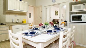 Un comedor con una mesa blanca dispuesta para cuatro personas, rodeada de sillas blancas. Se ve una cocina con microondas y armarios al fondo.