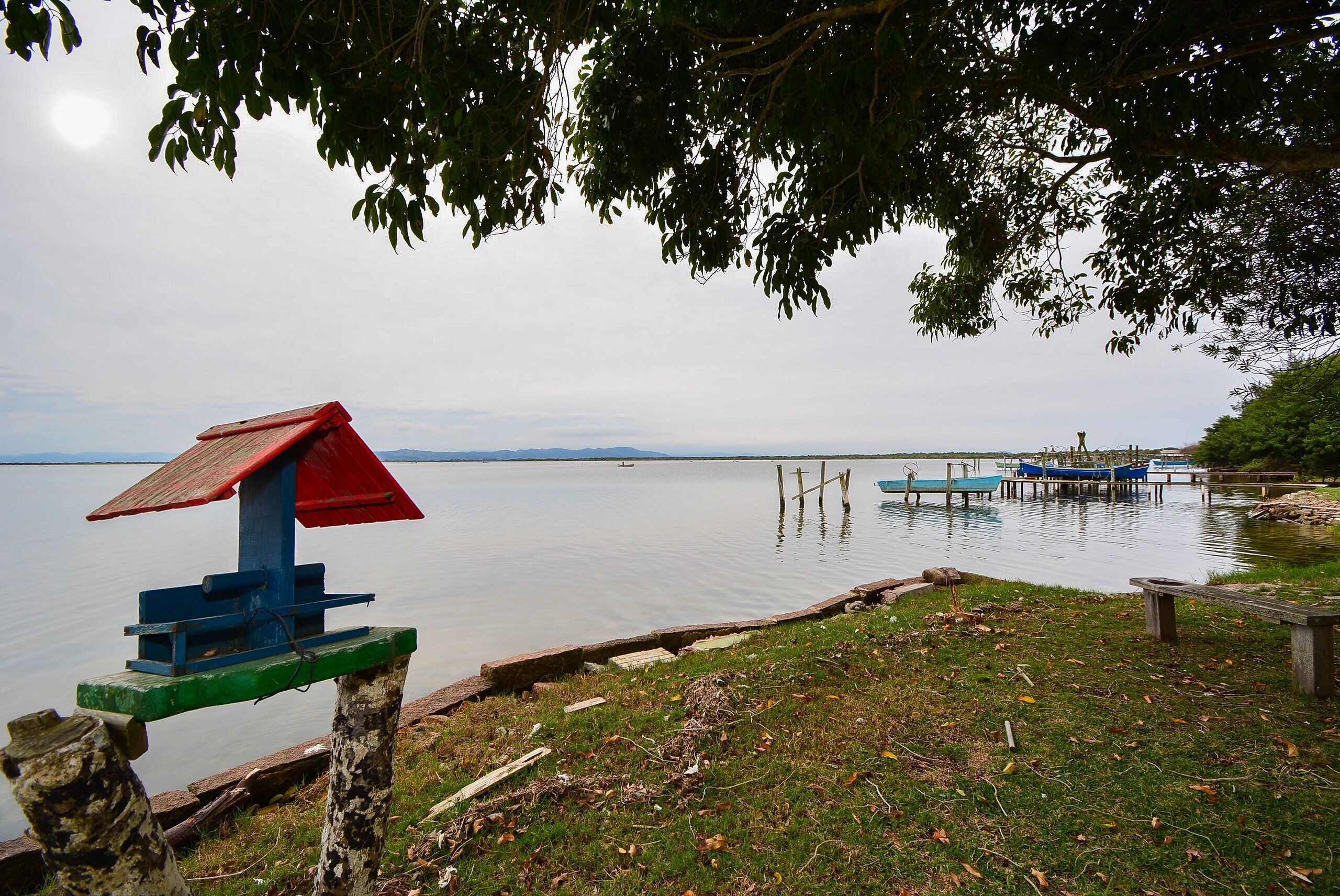 Escena junto al lago con un banco de madera, un comedero para pájaros y un muelle con barcos.