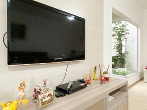 Una sala de estar moderna con un televisor de pantalla plana grande montado en la pared, un mueble de entretenimiento de madera con dispositivos electrónicos, y un ventanal con vistas a un árbol y un edificio.