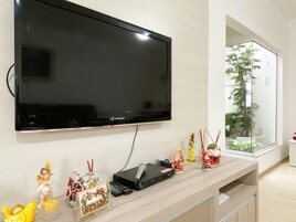 Uma sala de estar moderna com uma grande TV de tela plana na parede, um rack de madeira com dispositivos eletrônicos e uma janela ampla com vista para uma árvore e um edifício.