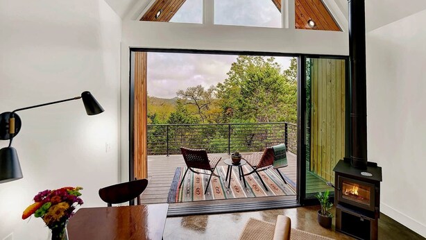 A modern living room with a large sliding glass door leading to an outdoor deck, a wood-burning stove, and a dining area with a table and chairs. - Wyldwood Modern Cabin - BRAND NEW! (Wimberley)