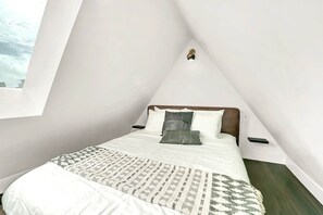 Ein Schlafzimmer mit schrägem Dach, einem Bett mit weißer Bettwäsche und einer gemusterten Tagesdecke, und Blick auf den Himmel durch ein Fenster.