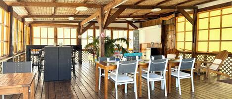 Een houten terras met een eethoek, voorzien van tafels en stoelen, een groot raam en een plafond met zichtbare balken.