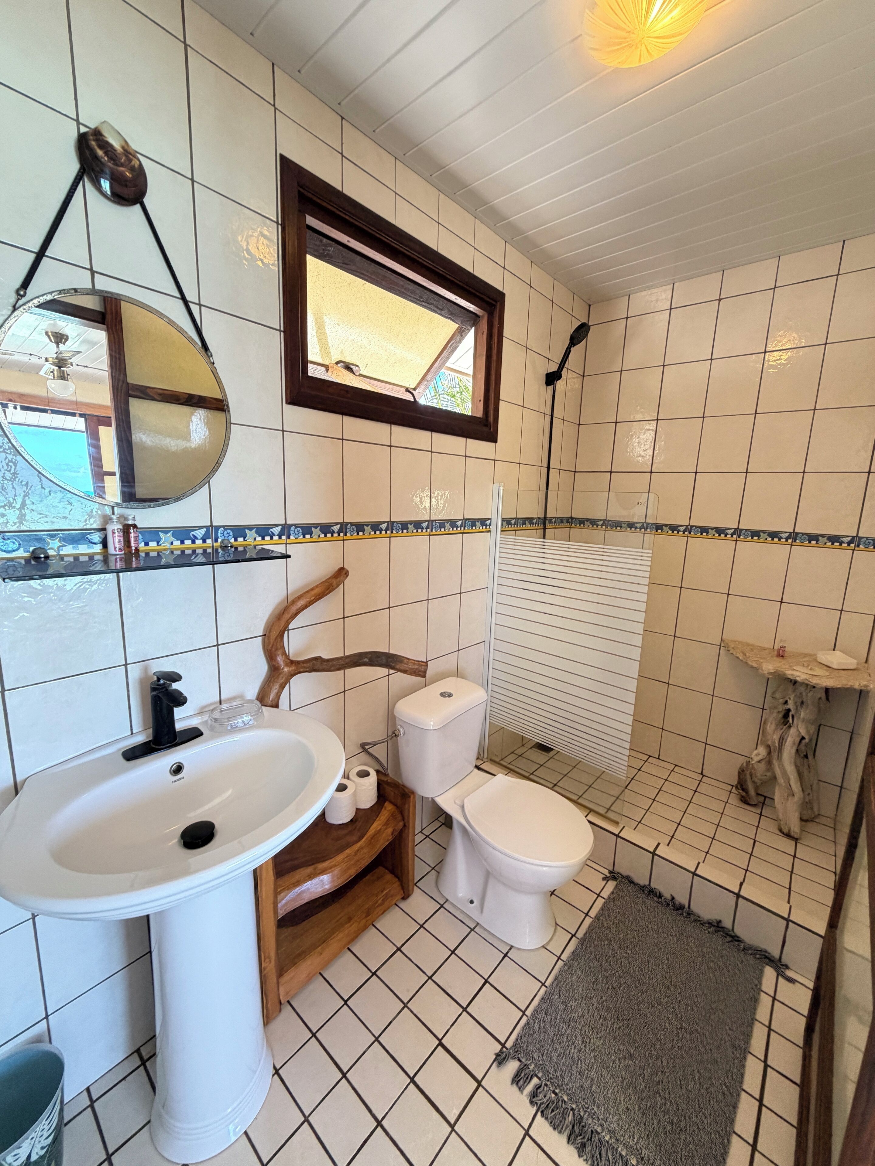A bathroom with a white sink, toilet, and shower area. There is a mirror above the sink, a window above the toilet, and a towel rack.
