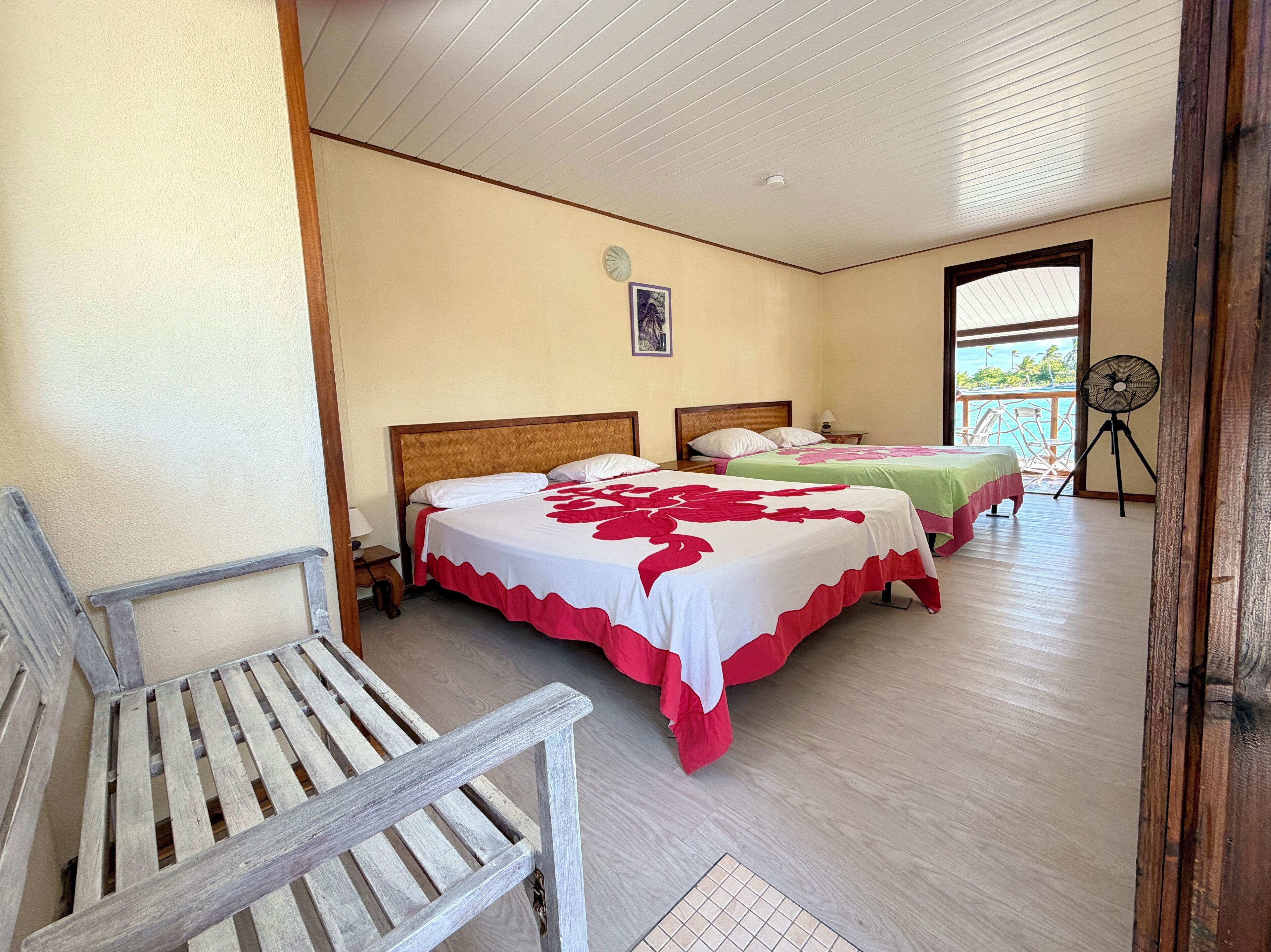Habitación de hotel con dos camas, una silla de madera, un ventilador y vista al mar.