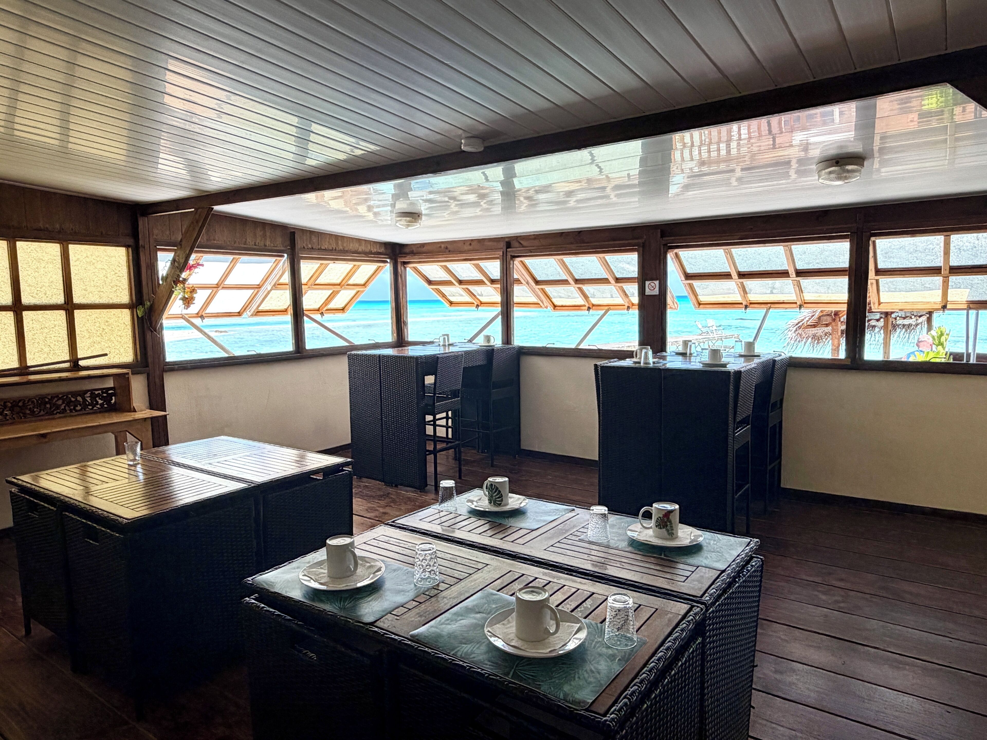 Een eetgedeelte met tafels en stoelen, grote ramen met uitzicht op de zee, en een houten plafond.