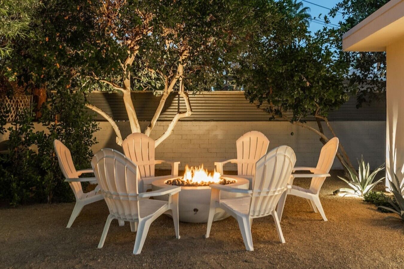 A patio with a fire pit and white chairs, surrounded by trees and a brick wall.