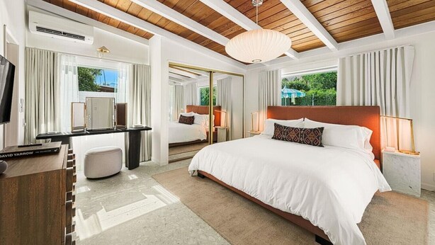 A modern hotel room with a large bed, a desk, a TV, and a view of greenery outside.