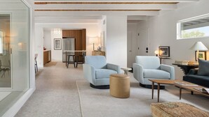 A modern living room with a high ceiling, a kitchen in the background, and a large window.