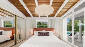 A spacious bedroom with a large bed, a wooden ceiling, and a view of a swimming pool.