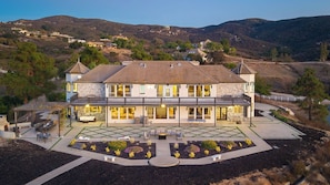 A large two-story house with a balcony, a patio with outdoor furniture, and a landscaped garden. - Hilltop Estate w/ Tennis Court, Pool, Sauna, Game Room, Theater, Panoramic Views Via de Oro Estate by Avantstay (Temecula)