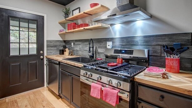 A modern kitchen with a stove, oven, and sink, wooden shelves above the counter, and a red utensil holder. - Park Avenue Retreat (Hot Springs)
