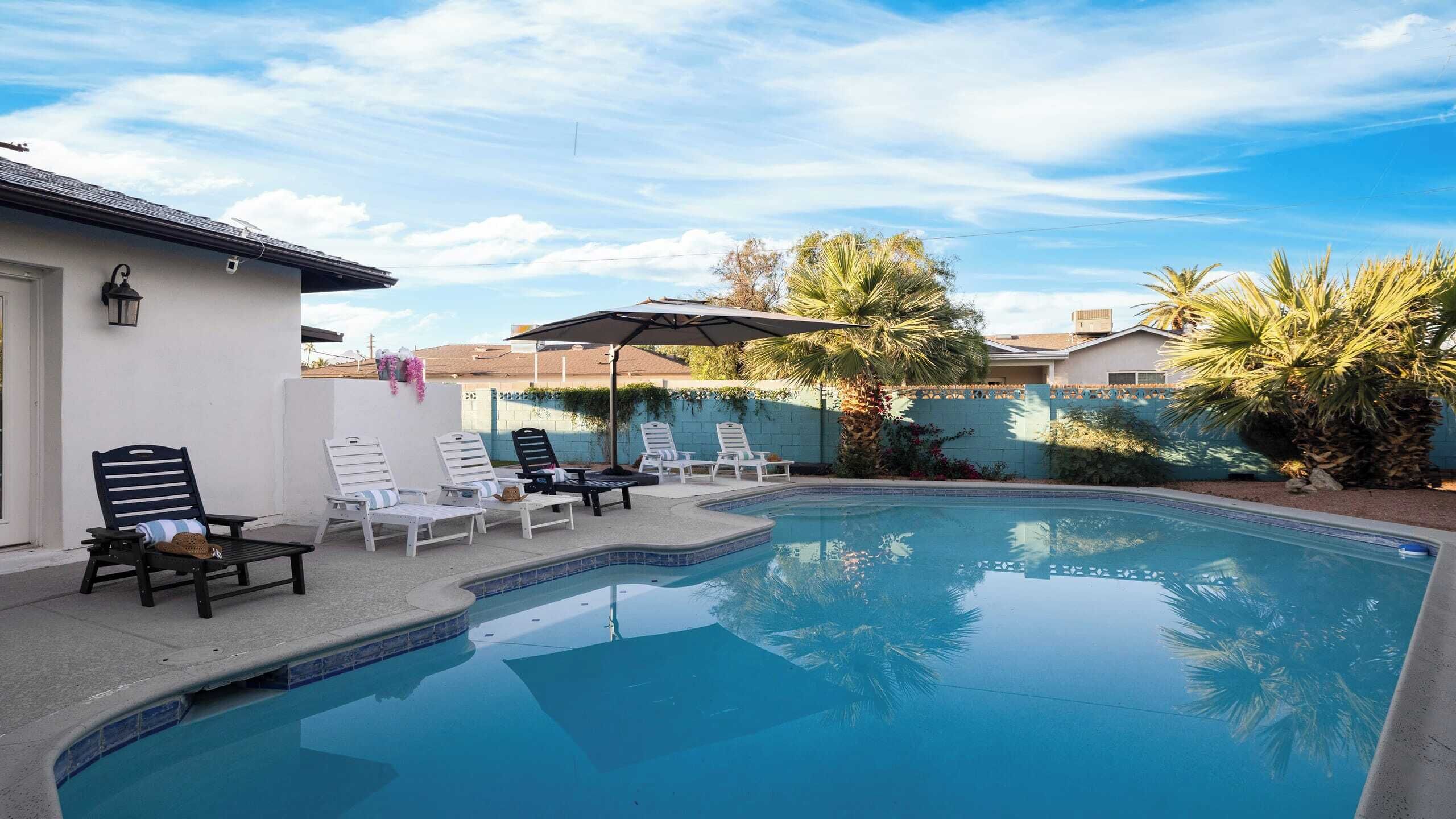 A swimming pool with lounge chairs and umbrellas, surrounded by palm trees and a white building.