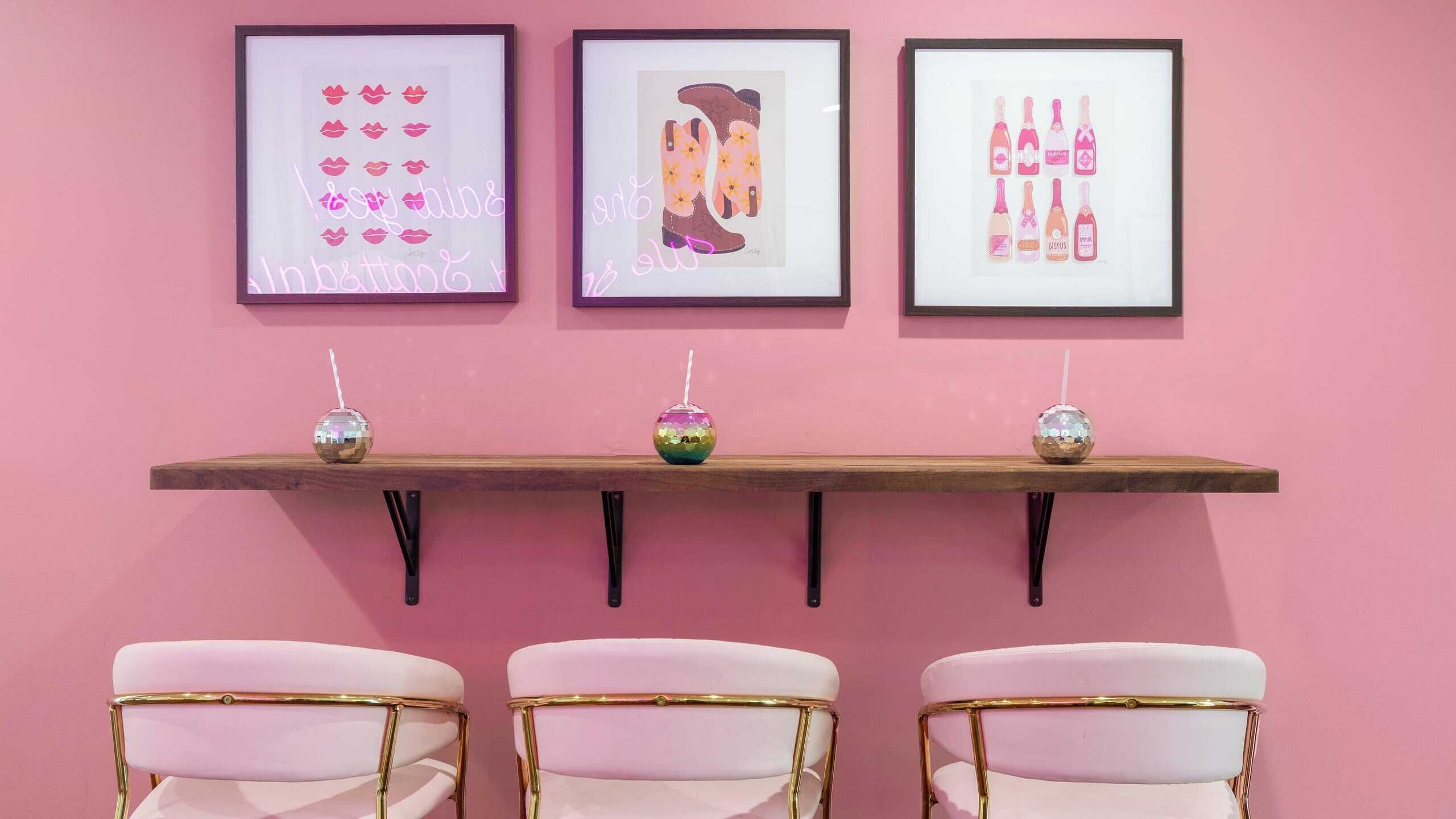 A pink wall with three framed artworks, a wooden shelf with three disco balls, and three pink chairs with gold accents.
