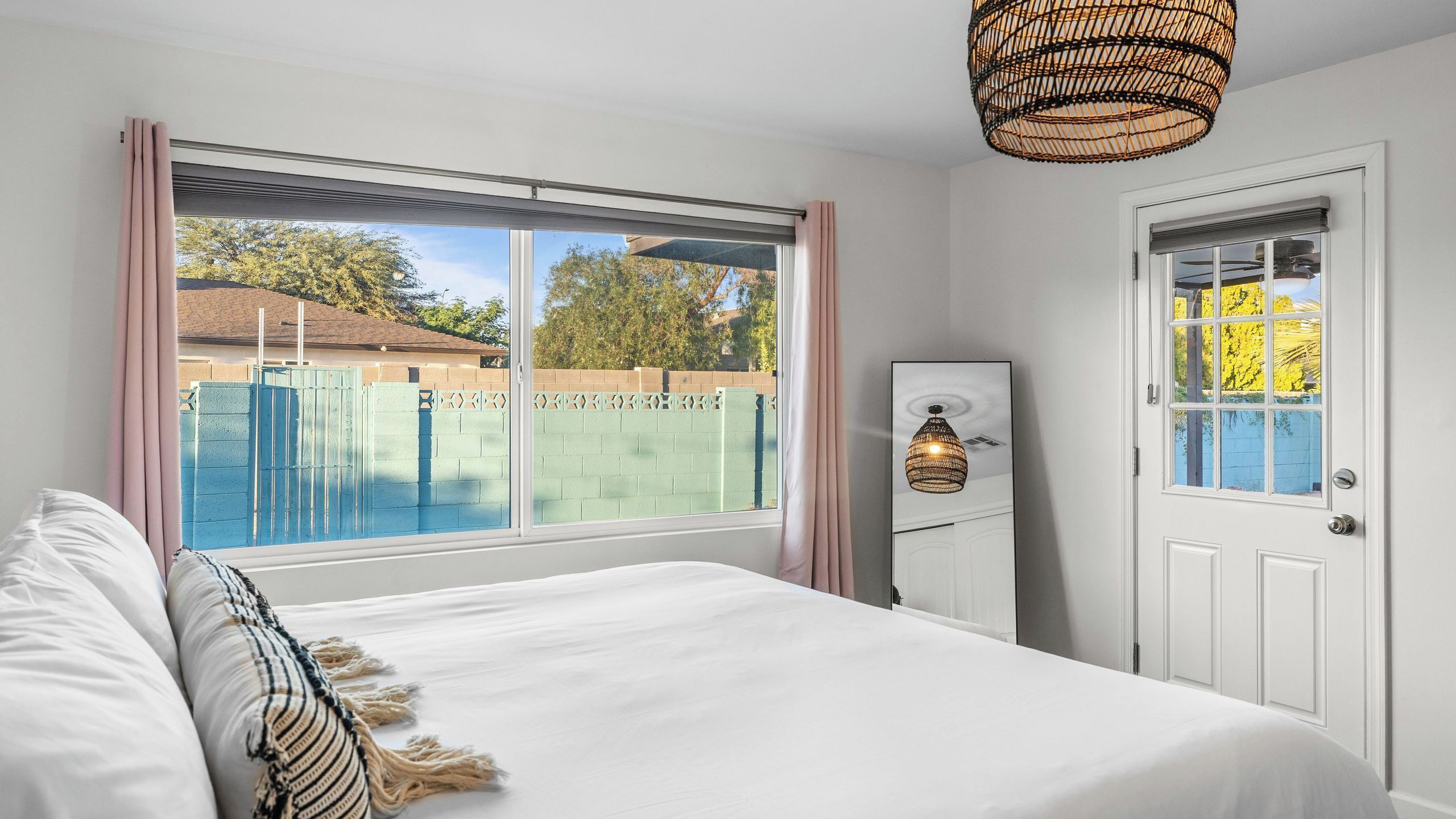 A bedroom with a large window, a bed with white bedding, a bedside table, and a door with a glass panel.